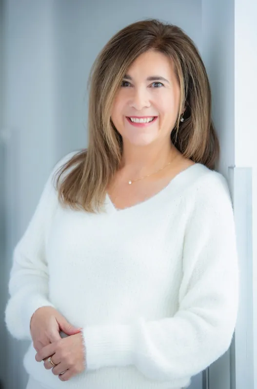 Johanne Côté souriante, appuyée contre un mur, en chandail blanc